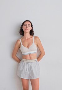 Model pozira uz ravni zid, obučena u bež bralet i prugaste šortseve, prenoseći opušten i ležeran stil.