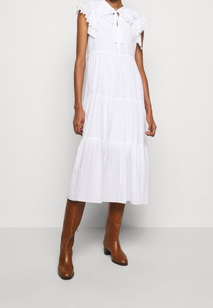 Biała sukienka midi bez rękawów z warstwową spódnicą i koronkowym kołnierzem zawiązanym na szyi, zestawiona z brązowymi skórzanymi kozakami do kolan.