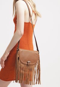 Žena v bezramenných oranžových pletených šatech, která nese hnědou koženou crossbody tašku se třásněmi na rameni.