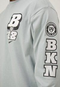 Langärmliges hellgraues Shirt aus weicher Baumwolle mit schwarzen Druckgrafiken, einschließlich "B12" und "BKN", sowie dem Logo der Brooklyn Nets auf dem Ärmel.