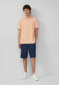 Helles Pfirsichfarbenes Polo-Shirt mit geripptem Kragen, kombiniert mit dunklen Jeansshorts. Das Modell trägt weiße Sneakers. Einfarbiger Hintergrund.