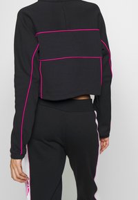 Svart cropped sweatshirt med långa ärmar, med rosa accentstygn över ryggen. Materialet verkar mjukt och strukturerat.