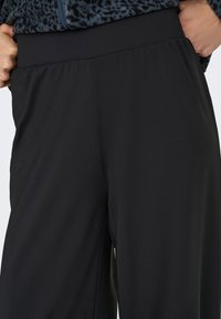 Μαύρο φαρδύ παντελόνι με ελαστική μέση που φοράει ένα άτομο που κρατά τη μέση, συνδυασμένο με σκούρο τοπ με σχέδια.