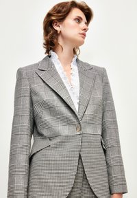 Harmaa ruutukuosisavereijakka, jossa on jämäkkä muotoilu, yksittäinen kultapainike, huippareunat ja rakennevalkoinen röyhelökauluspaita alla.