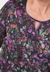 Bluse mit Blumenmuster in Schwarz, ausgestattet mit lila und grünen Blättern. Sie hat einen Schlitz-Ausschnitt und eine glatte, leichte Stoffstruktur.