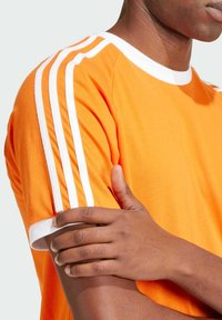 Camiseta naranja con ribete blanco y tres franjas blancas en cada hombro; material de algodón suave; diseño ajustado; mangas cortas.