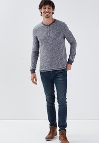 Maglietta henley grigia a maniche lunghe con scollo rotondo e pattina a tre bottoni, abbinata a jeans slim-fit blu scuro e stivali marroni.