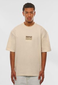 Beige oversized t-shirt van zachte katoen met korte mouwen, voorzien van herhaalde "DROPSIZE" tekst in verschillende bruintinten op de voorkant.
