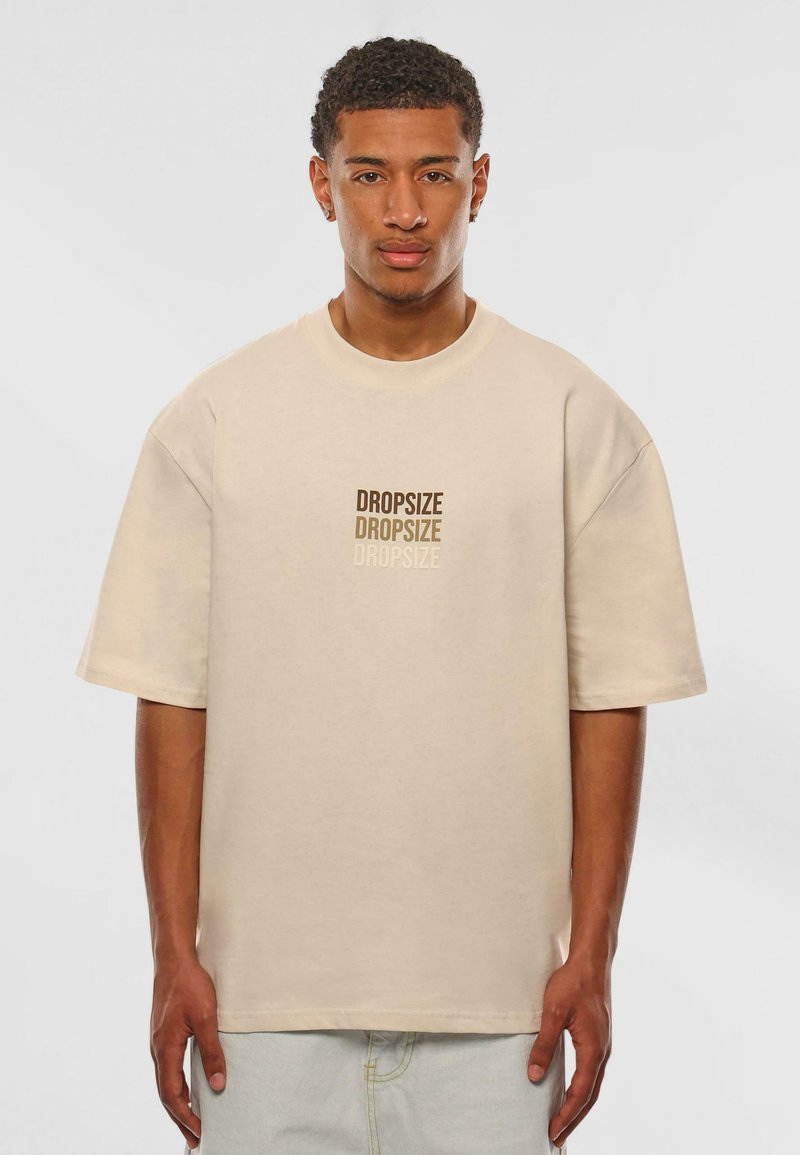 Beige oversized t-shirt van zachte katoen met korte mouwen, voorzien van herhaalde "DROPSIZE" tekst in verschillende bruintinten op de voorkant.
