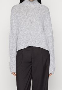 Lichtgrijze gebreide trui met een hoge nek, losse pasvorm en cropped lengte, gecombineerd met zwarte hoge taillebroeken. Zachte textuur.