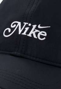 Μαύρο καπέλο κατασκευασμένο από υφασμάτινη υφή. Διαθέτει κεντητό λευκό λογότυπο "Nike" με στιλιζαρισμένα γράμματα, χωρίς ραφές και με καμπυλωτό γείσο.