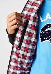 Main tenant une veste ouverte avec une doublure à carreaux rouges et blancs sur une chemise bleue avec un motif de crocodile noir et un texte blanc partiel.