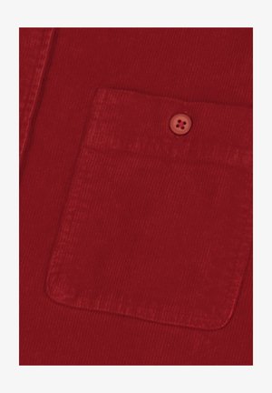 Primo piano di un tessuto in corduroy rosso che mostra una tasca quadrata con un bottone rosso abbinato cucito vicino al centro superiore.