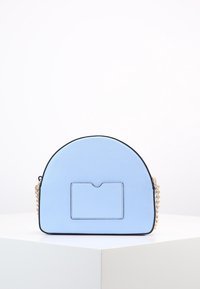 Vaaleansininen puolikuun muotoinen käsilaukku, jossa on etupuolen korttipussi ja kultaketju, esillä valkoisella jalustalla yksinkertaista taustaa vasten.