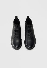 Botas de tobillo de cuero negro que presentan una puntera redondeada, cordones en el frente y cremallera lateral. Textura suave con diseño minimalista y tira de agarre.