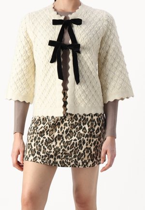 Kvinde iført en tekstureret cremefarvet cardigan med sorte fløjls sløjfer over en brun ribbet top og et mininederdel med leopardprint.