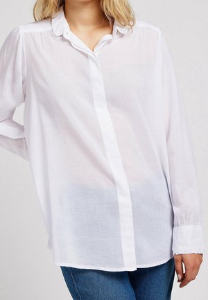 Kobieta nosząca luźną, białą bluzkę z długim rękawem i ukrytym listwą na guziki, połączoną z niebieskimi dżinsami, na jednolitym tle.