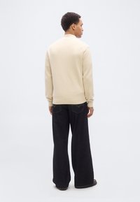 Άνδρας που φοράει μπεζ πουλόβερ και φαρδιά μαύρα τζιν, στέκεται γυρισμένος πλάτη σε ένα απλό λευκό φόντο.