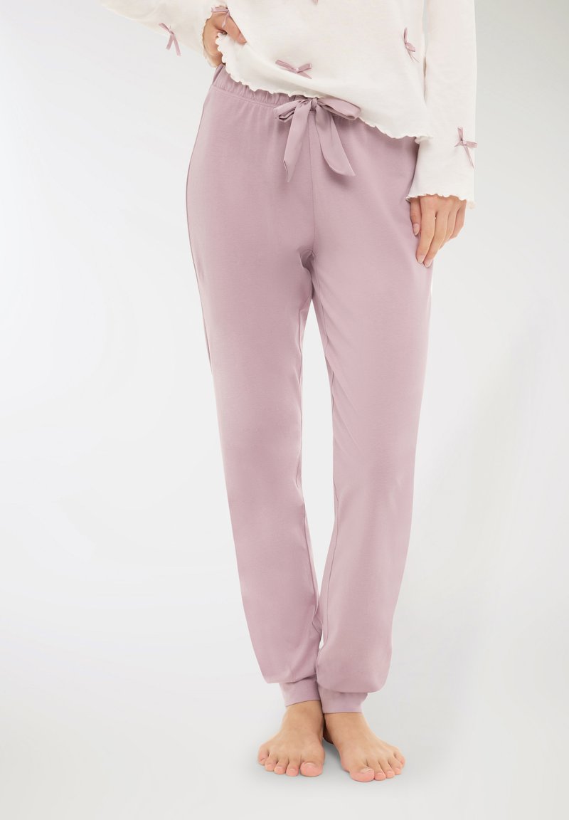 Donna che indossa pantaloni da pigiama in morbido tessuto lilla con un nastro legato in vita, abbinati a una maglietta bianca con piccoli dettagli a fiocco sulle maniche.