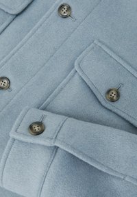 Világoskék gyapjúkabát nagy gombokkal, plüsszsebbel és gombos mandzsetta részlettel, közelről bemutatva.