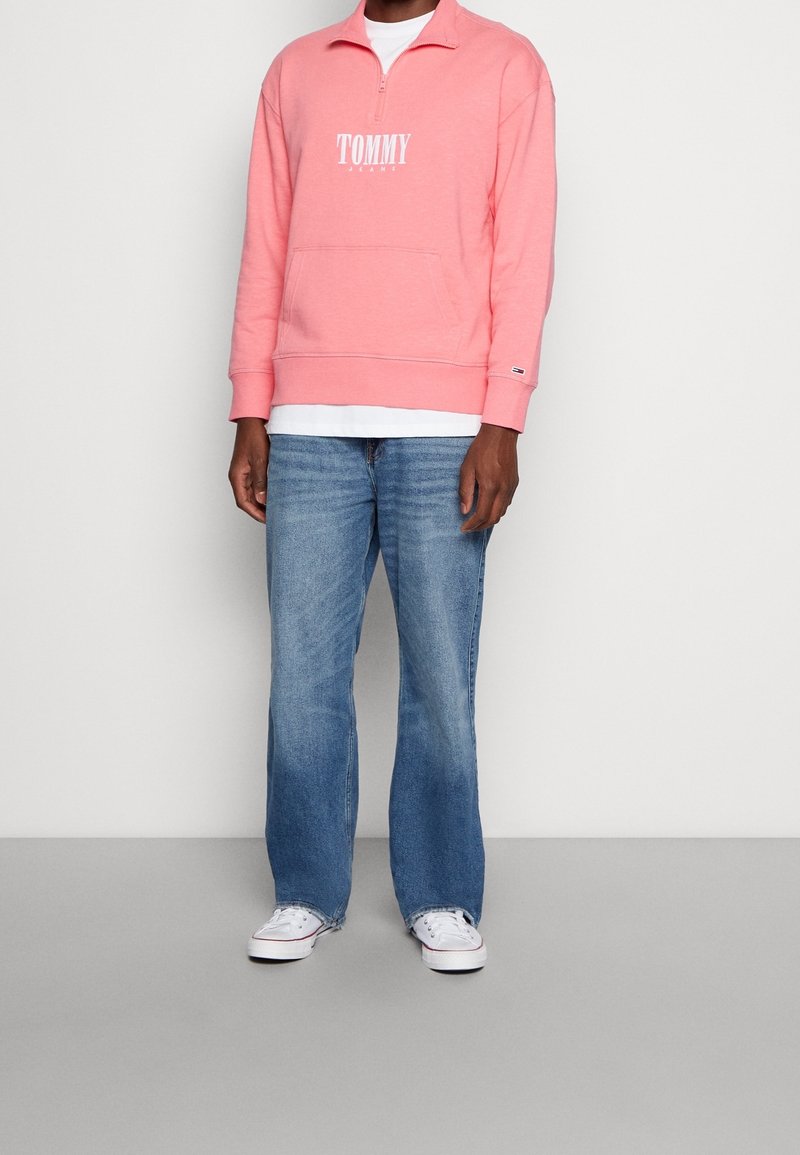 Personne portant un sweat rose avec une fermeture éclair au quart, avec le logo Tommy Jeans, un jean bleu droit, un t-shirt blanc en dessous et des baskets blanches.