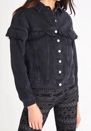 Sort denimjakke med hvide knapper og filtret vandret flæse hen over brystet og de øverste arme, båret med mønstrede sorte leggings.