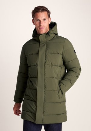 Man wearing green hooded puffer jacket with hands in pockets, standing against plain light background.