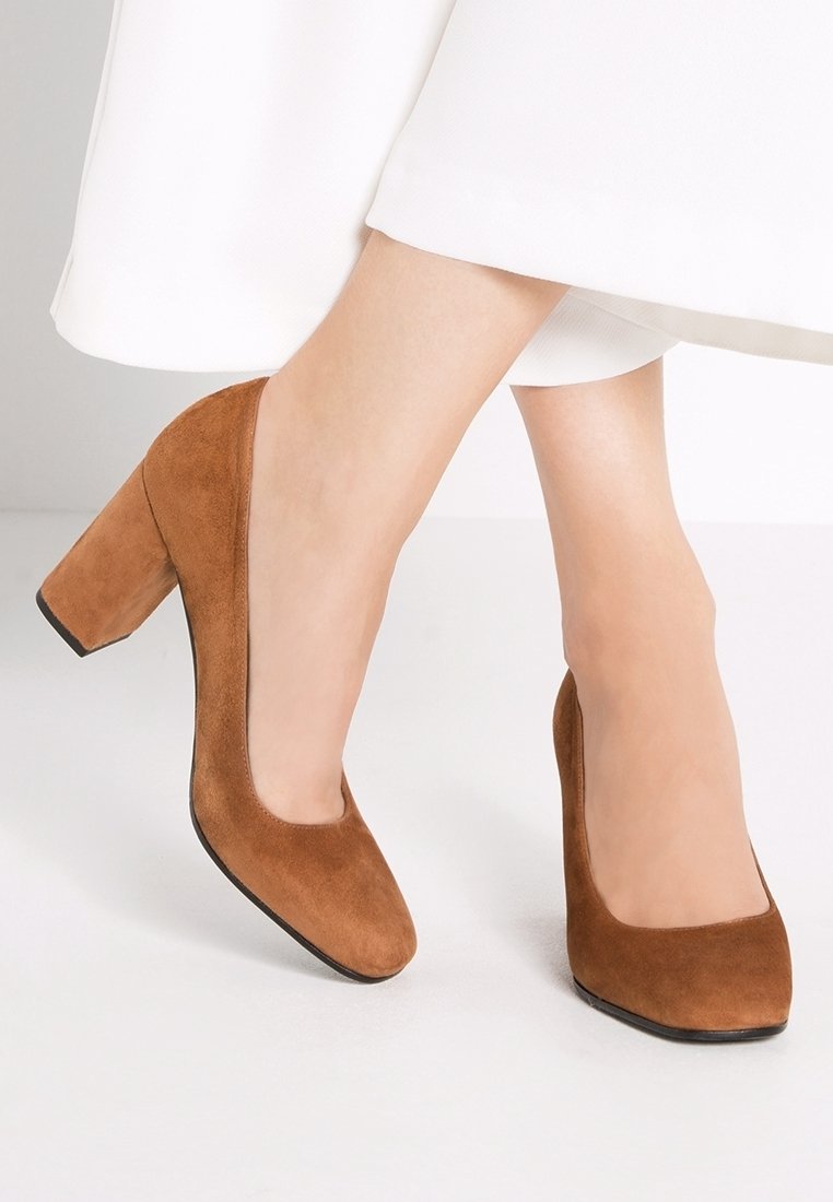 Des escarpins en daim marron avec un bout carré et un talon épais. La surface est lisse avec une texture subtile, se détachant sur un fond clair.