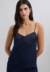 Marinblå klänning med V-ringning, tunna axelband och strukturerade prickdetaljer på den nedre delen, tillverkad av ett mjukt tyg.