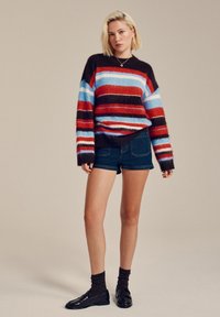 Multicolored striped sweater in brown, red, blue, and cream, paired with dark denim shorts and black loafers with textured socks.