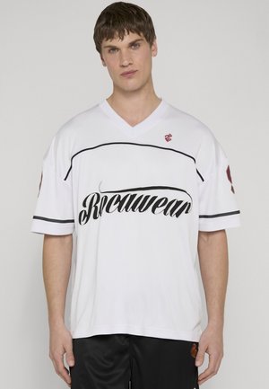 Mladić s kratkom smeđom kosom nosi široku bijelu majicu s crnim natpisom i crne šorceve na neutralnoj pozadini.