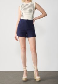 Navy high-waisted shorts with front pleats, paired with a white sleeveless top featuring black stripes. Model wearing beige strappy wedges.