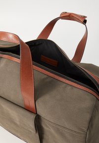 Khaki canvas duffel bag with brown leather straps, a smooth zipper, and a spacious black interior. Includes a small leather label inside.