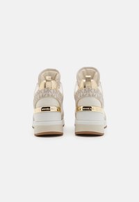 Sneaker con una toma superiore in tessuto beige caratterizzato da un motivo ripetitivo del logo, dettagli dorati sul tallone e una suola intermedia bianca con suola esterna marrone.