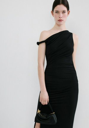 Woman with dark hair wearing a fitted one-shoulder black dress holding a small black handbag with gold chain detail, standing against a plain background.