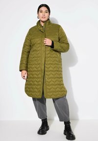 Cappotto lungo imbottito verde oliva con colletto alto, bottoni a scatto e polsini in maglia nera, abbinato a pantaloni grigi larghi e stivali neri chunky.