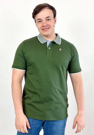 Jeune homme souriant, portant un polo vert avec un col gris et un jean bleu, se tenant devant un fond blanc.
