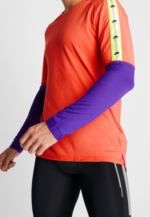 Homme portant un t-shirt orange à manches courtes avec une bande jaune fluo et des manches longues violettes en dessous, associé à un pantalon de sport noir.