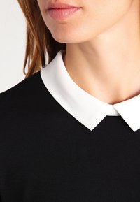 Svart topp med en vit, strukturerad krage. Tyget verkar slätt och något texturerat. Kragen skapar en skarp kontrast mot det svarta.