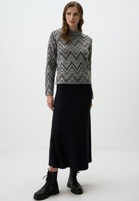 Pull en tricot à motif chevron gris, encolure ronde, associé à une longue jupe noire à côtes et des bottes de combat noires à lacets. Fond minimal.