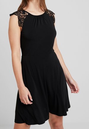 Frau trägt ein ärmelloses schwarzes Kleid mit Spitzendetails an den Schultern und einem ausgestellten Rock, steht vor einem schlichten grauen Hintergrund.