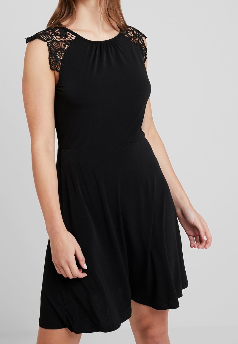 Femme portant une robe noire sans manches avec des détails en dentelle aux épaules et une jupe évasée, debout devant un fond gris uni.