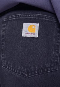 Mörk denimtyg nära bild med en vit etikett med en gul logotyp och texten "Carhartt" sydd på bakfickan.