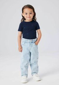 Jauns bērns ar garām matiem, ģērbts tumši zilā krekliņā ar sirds rakstiem, gaiši zilos džinsos ar maziem ziedu rakstiem un baltiem sporta apaviem.