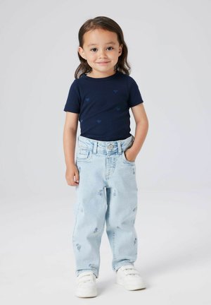 Ungdommelig barn med langt hår som har på seg en marineblå skjorte med hjerte-mønster, lyseblå jeans med små blomstermotiver, og hvite sneakers.