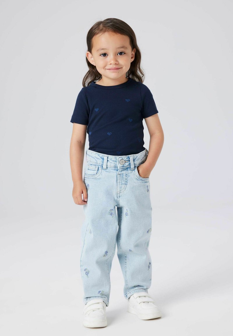 Jauns bērns ar garām matiem, ģērbts tumši zilā krekliņā ar sirds rakstiem, gaiši zilos džinsos ar maziem ziedu rakstiem un baltiem sporta apaviem.