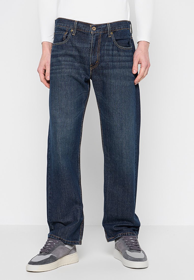 Homme portant un jean droit bleu marine et des baskets grises, se tenant devant un fond blanc uni.