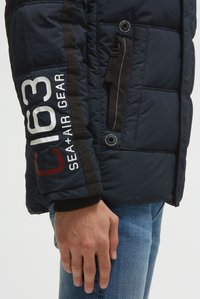 Hand van een man langs zijn zijde, gekleed in een marineblauw gewatteerd jack met de tekst "C 193 SEA + AIR GEAR" op de mouw en blauwe spijkerbroek tegen een witte achtergrond.