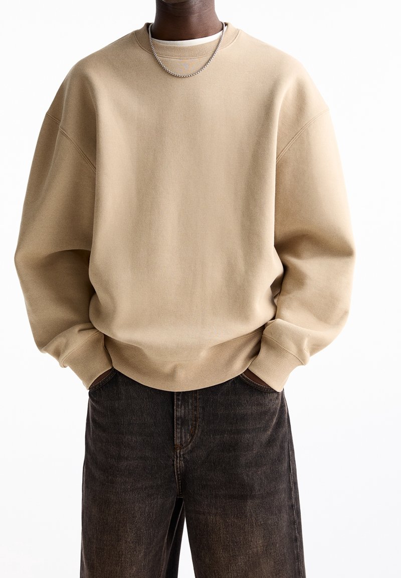 Beige ylipitkä collegepaita, jossa on ribbireunukset kauluksessa ja hihansuissa, yhdistettynä tummansinisisiin farkkuihin. Kangas näyttää pehmeältä ja sileältä.