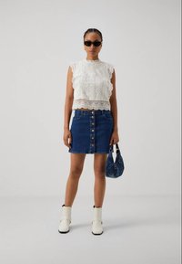 Weißes Spitzencrop-Top, Denim-Skirt mit Knopfleiste, weiße Knöchelstiefel und handgefertigte blaue Handtasche mit Muster. Dunkle Sonnenbrillen runden den Look ab.